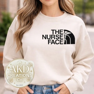 Peut inclure: Sweat-shirt crème avec l'inscription noire "THE NURSE FACE" et un symbole médical. Le sweat-shirt est associé à un jean bleu. Un logo circulaire avec le texte "AKD Creations Co. Est. 2023" est visible.