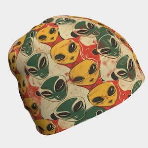 May include: A beige beanie with a repeating pattern of green, yellow, and red alien faces. The beanie has a distressed, vintage look.