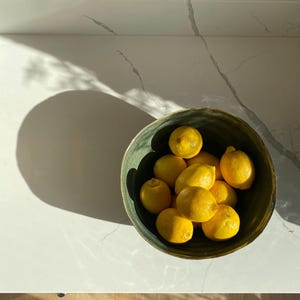May include: A green ceramic bowl filled with yellow lemons. The bowl is on a white countertop.