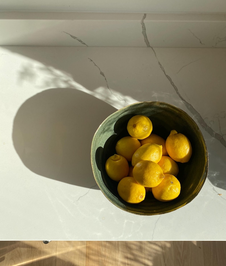 May include: A green ceramic bowl filled with yellow lemons. The bowl is on a white countertop.
