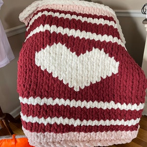 May include: A chunky knit throw blanket with a large white heart in the center. The blanket is striped with burgundy and white, and has a pink border.