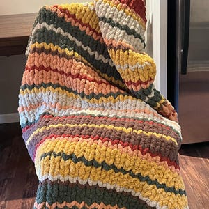 Pode incluir: Uma manta de malha grossa com um padrão listrado em tons de vermelho, laranja, amarelo, verde, marrom e branco.