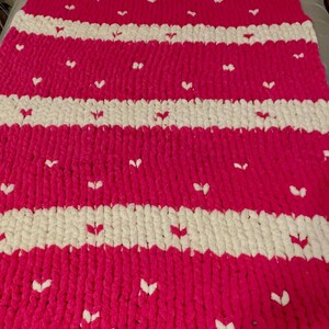 May include: A handmade, chunky knit blanket in a vibrant fuchsia pink. The blanket features horizontal stripes of white and is dotted with small white heart-shaped accents. The cozy throw is perfect for adding warmth and style to any room.