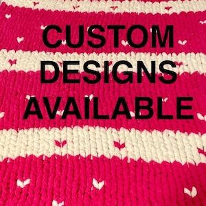 May include: A pink and white striped knitted blanket with white hearts scattered throughout. The blanket has the text "CUSTOM DESIGNS AVAILABLE" in black letters.