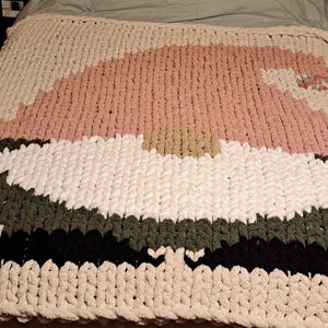 May include: A chunky knit blanket featuring a gnome design. The blanket uses a color palette of cream, pink, white, green, and black. The gnome's hat is pink, and the beard is white. The blanket is on a bed.