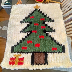 Pode incluir: Uma manta de crochê branca com um design de árvore de Natal verde, preto e marrom. A árvore tem enfeites vermelhos e uma estrela dourada no topo. Há dois presentes dourados e vermelhos na parte inferior da árvore.