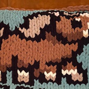 May include: A handmade, chunky knit blanket featuring a pixelated elephant design. The blanket is primarily light blue with a brown, black, and white elephant. The edges are finished with a brown border, creating a cozy and decorative item.