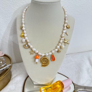 May include: A pearl necklace with gold charms, including a seashell, starfish, and mushroom. The necklace is displayed on a mannequin. A bottle of perfume is visible in the foreground.