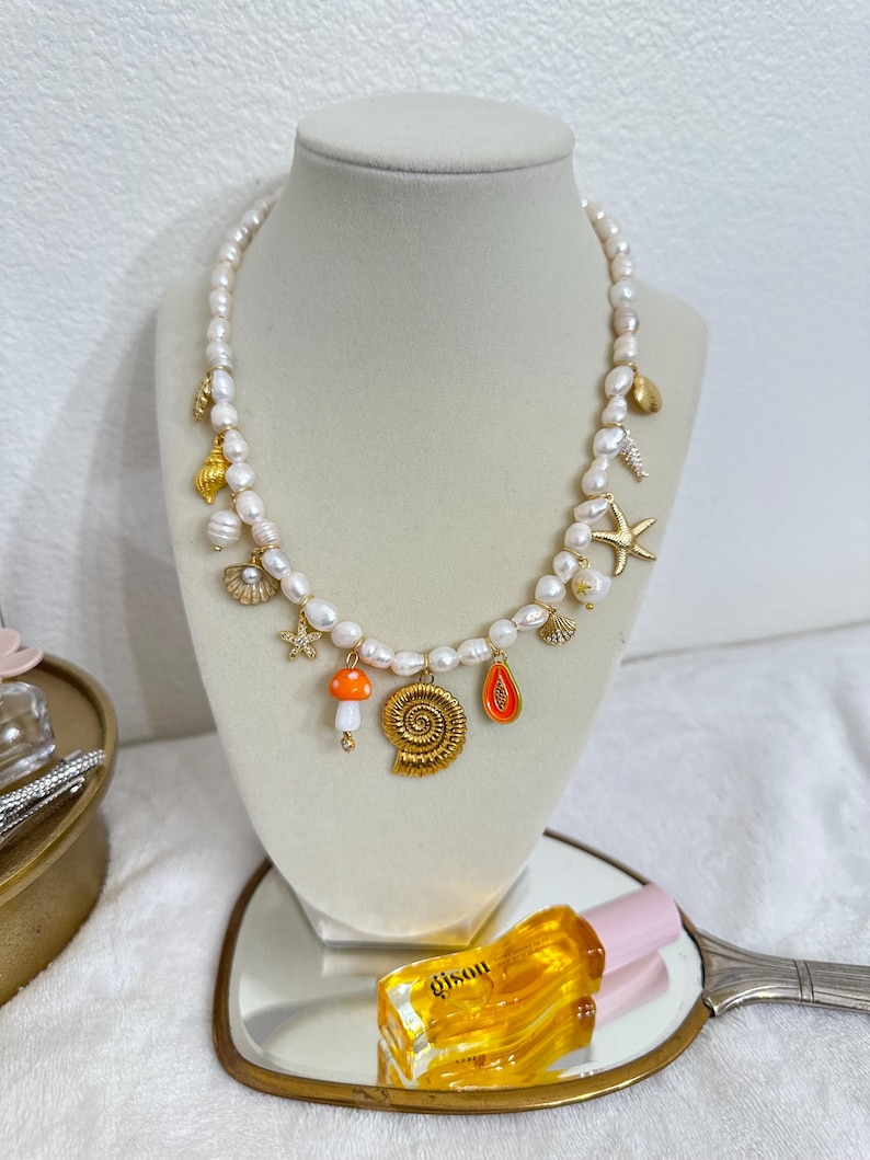 May include: A pearl necklace with gold charms, including a seashell, starfish, and mushroom. The necklace is displayed on a mannequin. A bottle of perfume is visible in the foreground.