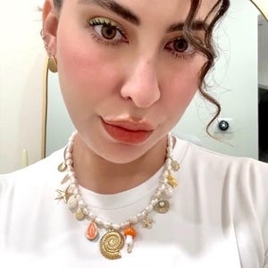 May include: A white pearl necklace with gold charms, including a seashell, starfish, and mushroom. The necklace is worn with a white t-shirt. Gold earrings are also visible.