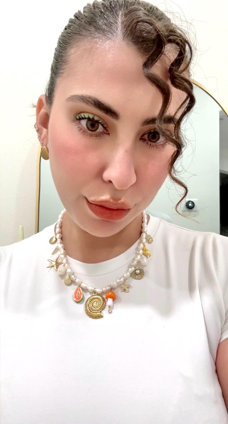 May include: A white pearl necklace with gold charms, including a seashell, starfish, and mushroom. The necklace is worn with a white t-shirt. Gold earrings are also visible.