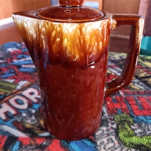 May include: A ceramic pitcher with a lid, featuring a glossy dark brown base that transitions into a lighter cream and tan drip glaze near the top rim. The pitcher has a sturdy handle, a pointed pouring spout, and a round knob on the lid.