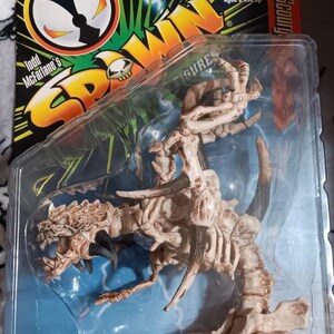 May include: A Spawn action figure in original packaging featuring a skeletal creature with beige bones and dark horns. The packaging includes the text Spawn, Todd McFarlane's, Ages 5 and Up, and Ribcage Holds Little Fiend, with a small blue figure.