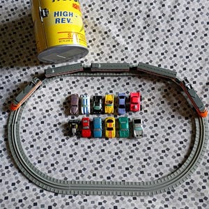 May include: A yellow cylindrical container labeled Secret Auto Supplies Motor High Rev with a set of grey plastic train tracks arranged in an oval. Inside the tracks are twelve small toy cars in various colors including red, blue, green, and yellow.
