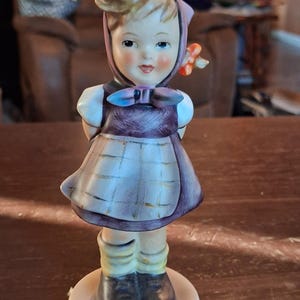 May include: A ceramic figurine of a child wearing a purple headscarf, a purple dress with a white checkered apron, yellow socks, and dark brown shoes. The figure has blonde hair, rosy cheeks, and stands on a circular base with a glossy finish.