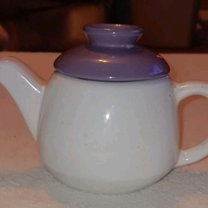 May include: A small ceramic teapot featuring a glossy white rounded body with subtle yellow speckles, a curved spout, and a matching handle. The teapot is topped with a smooth, solid lavender-colored lid that has a small knob handle at the center.