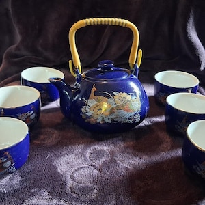 May include: A dark blue ceramic tea set consisting of a teapot with a yellow bamboo-style handle and five matching tea cups. The teapot and cups feature a detailed gold, orange, and green peacock and floral design with gold-trimmed rims.