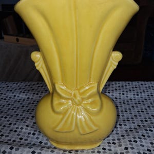 May include: A bright yellow ceramic vase featuring a wide, flared opening and a rounded base. The exterior is decorated with a molded bow detail at the center, flanked by vertical ridges that extend upward toward the rim of the vessel.