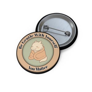 Könnte beinhalten: Ein runder Button mit schwarzem Rand und silberner Nadel. Der Button zeigt einen Cartoon-Drachen, der in eine Decke gehüllt ist, mit dem Text "Be Gentle With Yourself" und "You Matter" in einem kreisförmigen Design.