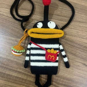 May include: A whimsical, handmade crochet crossbody bag in the shape of a cartoon character. The bag is primarily black with white stripes, a yellow mouth, and a red strap. It features a small burger charm and a red pocket with a fries logo.
