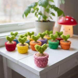 May include: A collection of colorful, crocheted heart-shaped plants in small pots. The pots are in various colors, including red, yellow, orange, green, and purple. The plants sit on a white surface, with a larger plant in the background.