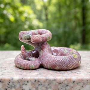 Może przedstawiać: Mała, zwinięta ceramiczna figurka grzechotnika o teksturowanym, błyszczącym wykończeniu w odcieniach przybrudzonego różu, ozdobiona rozproszonymi jasnymi, zielonymi i żółtymi plamkami na ciele, z ogonem uniesionym w charakterystycznej pozycji.
