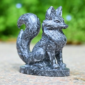 May include: A detailed, gray and white fox figurine. The fox is in a seated position with its tail curled. The sculpture has a patterned design and is set on a rectangular base. The background is blurred green.