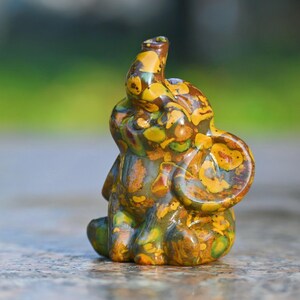 Puede incluir: Pequeña figura decorativa de elefante en tonos amarillos, marrones y verdes. El elefante está sentado con la trompa levantada y las orejas visibles. La figura está hecha de un material moteado.