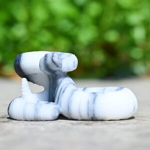 May include: A white and grey marble-effect snake ornament. The sculpture features a rounded base, a raised head, and a curled tail. The snake's body is white with grey streaks, and it rests on a wooden surface.