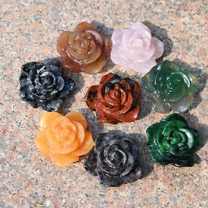 May include: A collection of carved stone roses in various colors, including orange, pink, green, and black. The roses are arranged in a circular pattern, showcasing their detailed petal designs. The gemstones are displayed on a speckled, neutral-toned surface.