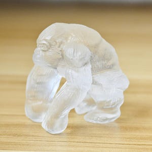 May include: A clear, translucent gorilla sculpture. The gorilla is depicted in a seated position, with its arms and legs visible. The sculpture has a smooth, polished appearance and is set against a light brown background.