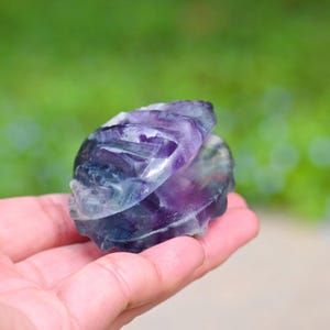 Op de afbeelding: Een gebeeldhouwde fluorietkristallen sculptuur in tinten paars, blauw en wit wordt in een hand gehouden. De sculptuur heeft een afgeronde vorm met gebeeldhouwde details. De achtergrond is een wazig groen.