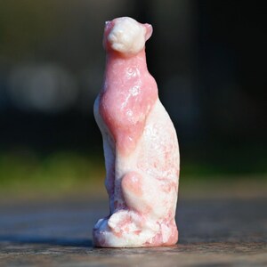 May include: A pink and white carved stone sculpture of a seated cat. The cat is depicted with its head up, and the pink color is concentrated on the upper body. The sculpture is detailed and has a smooth finish.