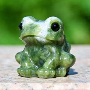 Könnte beinhalten: Eine kleine, aus Stein geschnitzte Froschfigur mit einer polierten, gesprenkelten Oberfläche in Dunkelgrün und Weiß. Der Frosch hat große, glatte, schwarze runde Augen und einen abgerundeten Körper mit deutlich erkennbaren Beindetails.