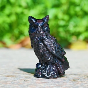 May include: A dark purple owl ornament with textured feather detailing. The owl is depicted perched on a small base. The background features blurred green foliage, accentuating the owl's form.