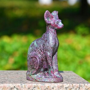May include: A seated cat sculpture with a Sphinx-like appearance. The stone cat statue is a mix of purple, green, and red hues. The sculpture is displayed on a marble surface with a blurred green background.