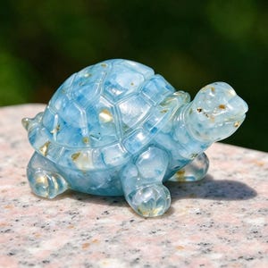 May include: A small, translucent light blue turtle figurine crafted from resin. The shell and body feature a smooth, polished surface with embedded gold-colored flecks throughout the material, creating a shimmering effect within the light blue base.
