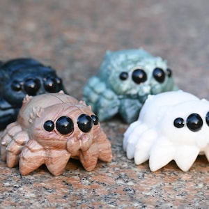 May include: Four small, detailed spider figurines in varying colors: dark blue, green, tan, and white. Each has large, black eyes and a textured body, resembling jumping spiders. The figurines are arranged on a speckled surface.
