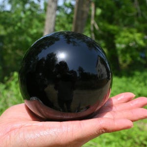 Op de afbeelding: Een grote, zwarte obsidiaankogel, gepolijst tot een hoge glans, wordt in de hand van een persoon gehouden. De bol reflecteert de omgeving en creëert een spiegelachtig effect.