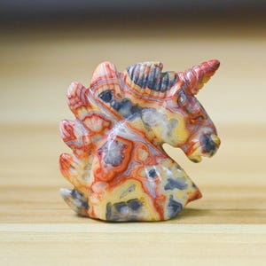 May include: A carved stone unicorn figurine. The sculpture features a detailed mane and horn, with a color palette of red, yellow, gray, and white. The figurine is set against a light brown background.