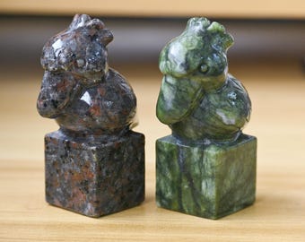 May include: Two stone sculptures of birds, one in shades of brown and the other in green, each perched atop a square base. The bird figures have detailed features and a polished finish, showcasing the natural patterns of the stone.