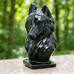 Puede incluir: Una escultura negra pulida de la cabeza y el cuello de un lobo, con texturas de pelaje detalladas y orejas puntiagudas. La figura está montada sobre una base cuadrada y presenta una superficie lisa y reflectante.