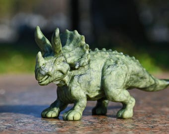Op de afbeelding: Een groene Triceratops-figuur met een textuur. De dinosaurus heeft drie hoorns, een kraag en een korte staart. De figuur staat op een bruin oppervlak.
