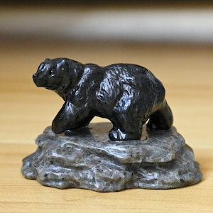 May include: A small, black bear figurine carved from stone. The bear is depicted in a walking pose, atop a gray, textured rock base. The sculpture has a polished finish, and the details of the bear's fur are visible.