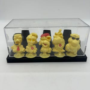 May include: A set of five yellow figurines displayed in a clear plastic case. Each figurine has a unique design, with some wearing hats or sunglasses. The figurines are mounted on a black base.