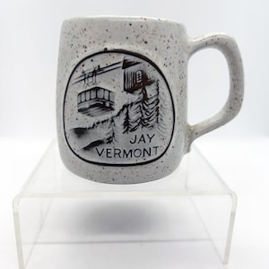 May include: Speckled ceramic mug featuring a black and white illustration of a mountain scene and the text "JAY VERMONT". The mug has a curved handle and is displayed on a clear acrylic stand.