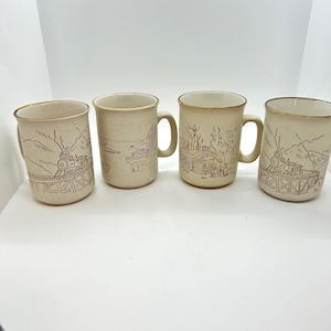 Peut inclure: Quatre tasses en céramique beige avec des dessins au trait marron d'un train, d'une cabane et d'un paysage lacustre. Chaque tasse a un bord doré et une anse. Les tasses sont de taille et de forme similaires.