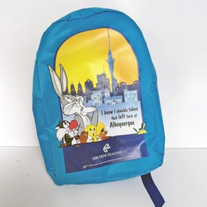 Könnte beinhalten: Blauer Rucksack mit Looney Tunes-Motiv, darunter Bugs Bunny, Tweety und andere Charaktere. Die Grafik zeigt eine Skyline und den Text "I knew I shoulda taken that left turn at Albuquerque." Er hat einen blauen Riemen.
