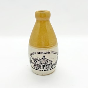 Könnte beinhalten: Kleine, vintage Keramikflasche mit braunem Oberteil und weißem Boden. Der Boden zeigt eine Schwarz-Weiß-Illustration eines Gebäudes mit dem Text "Upper Canada Village" und "Canada".