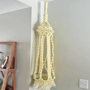 May include: Cream-colored macrame plant hanger suspended from a ceiling hook. The handmade hanger features a woven design with a circular top, a cylindrical body, and a fringed bottom. The plant holder is designed to hold a potted plant.